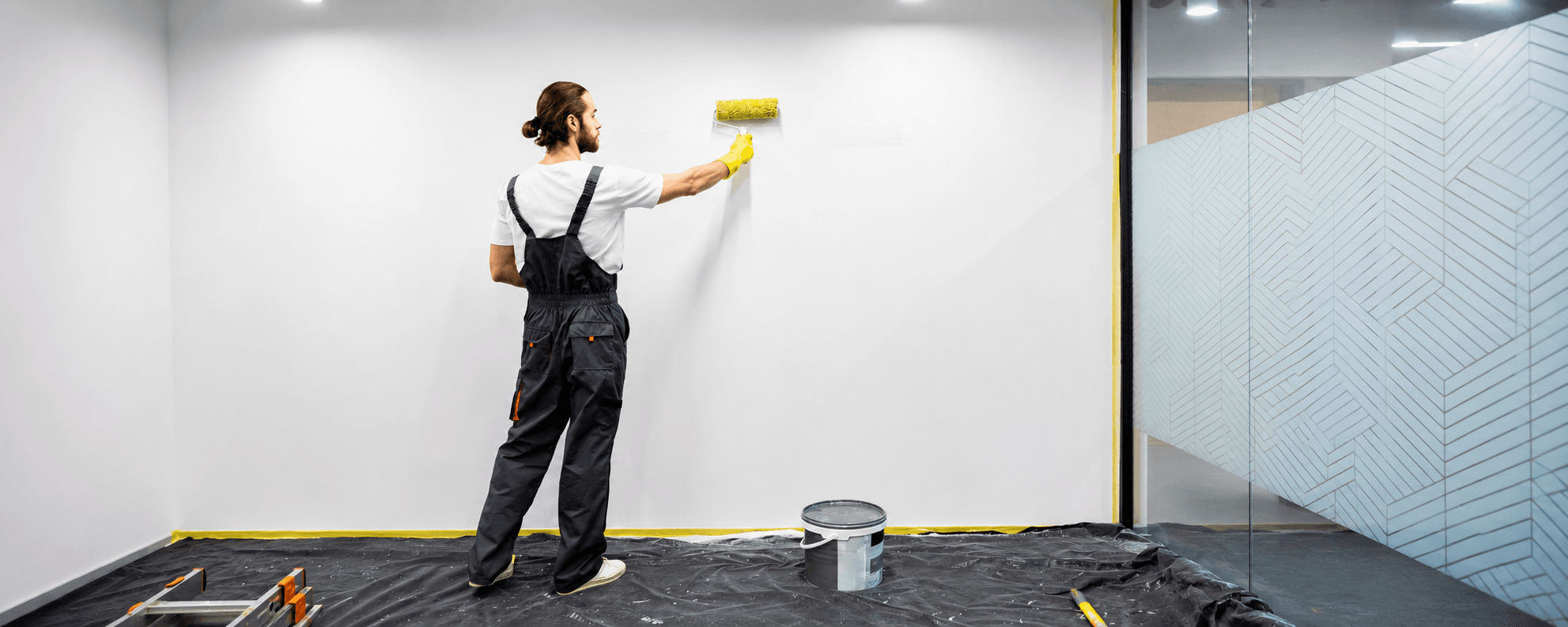 Decorator applying clear dry erase wall paint to a white wall with a roller, with masking tape, floor protection sheets and a paint tub in place.
