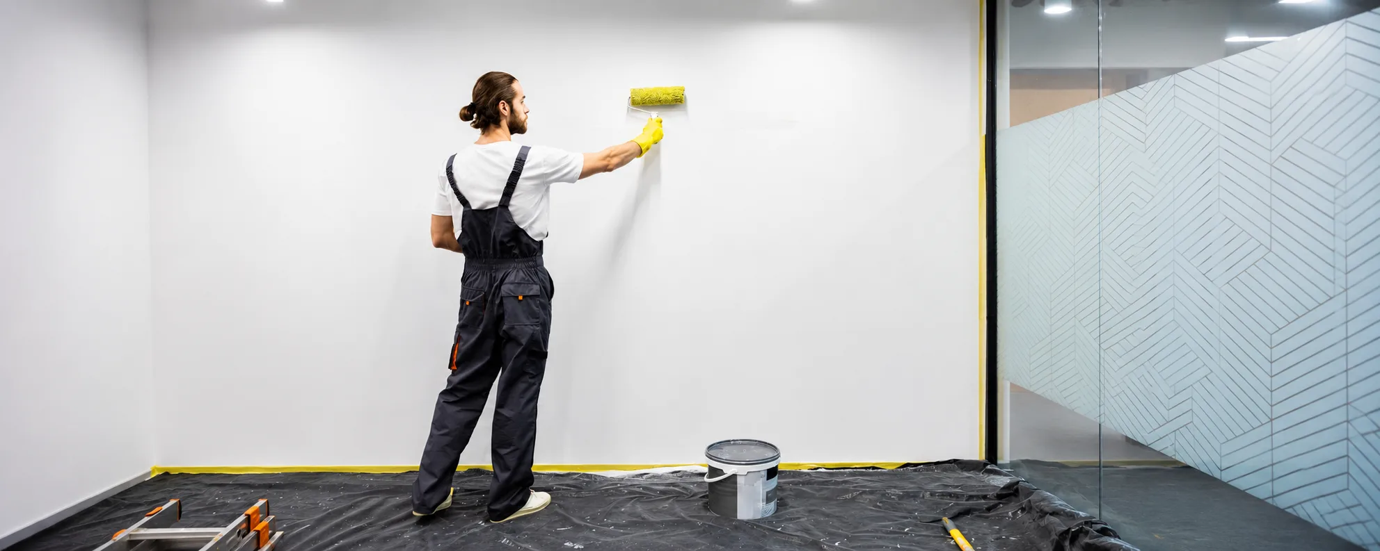 Picture7 Decorator applying clear dry erase wall paint to a white wall with a roller, with masking tape, floor protection sheets and a paint tub in place.